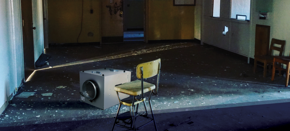 An abandoned room with peeling floors and scattered debris, illuminated by sunlight through a doorway. A HEPA air scrubber sits beside an old chair, highlighting restoration efforts in a damaged space