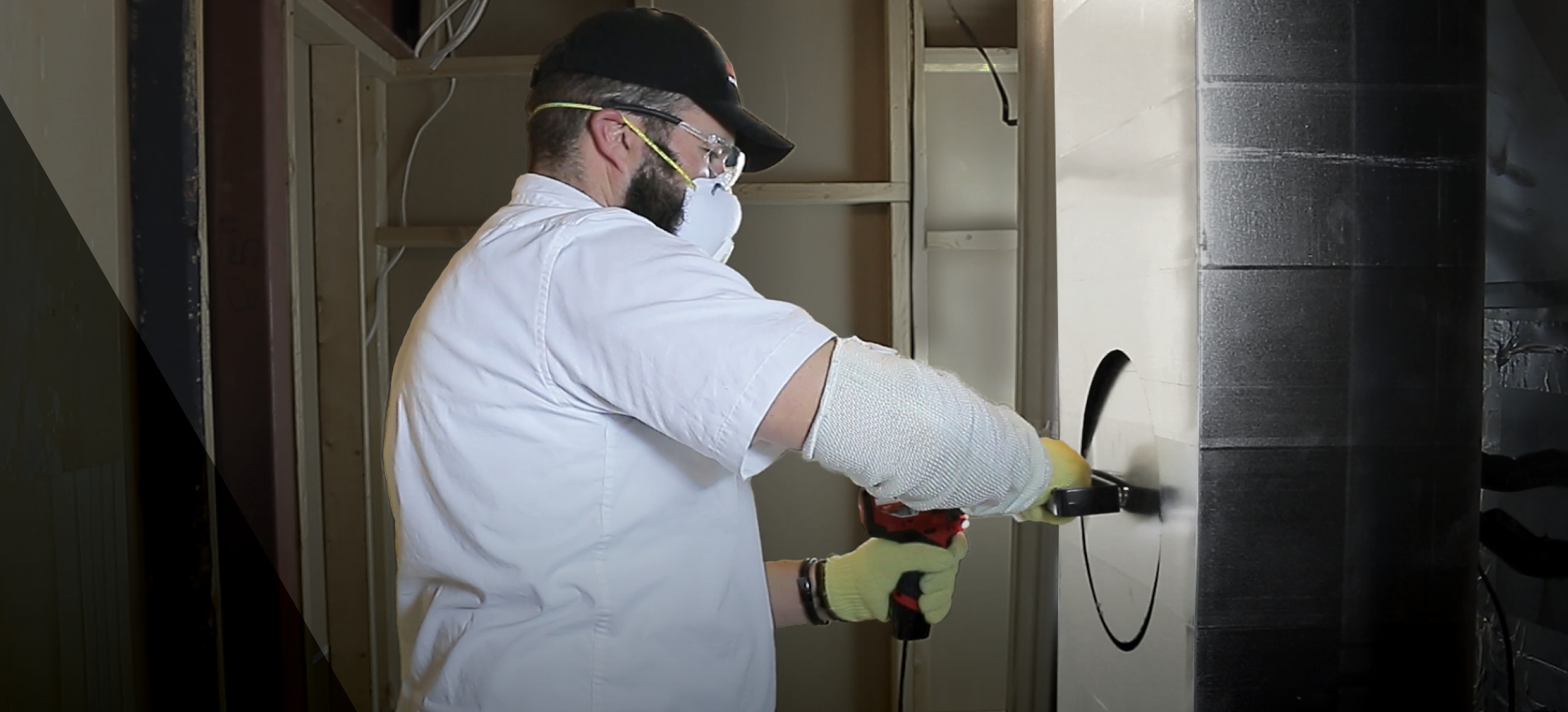 Contractor cutting a circular opening into an air containment barrier during restoration setup. Wearing protective gear including gloves, safety glasses, and a respirator mask, the worker prepares a negative air system for dust and mold control in a const