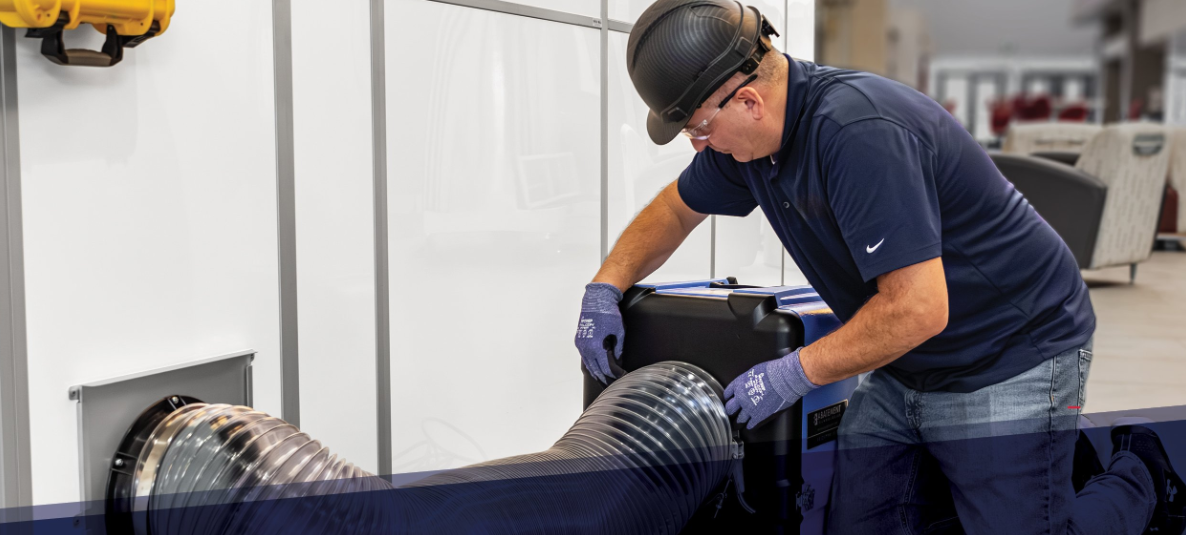 Technician setting up a HEPA air filtration system using flexible ducting, connected to a temporary containment barrier (SHIELD WALL) in a healthcare or commercial facility setting.